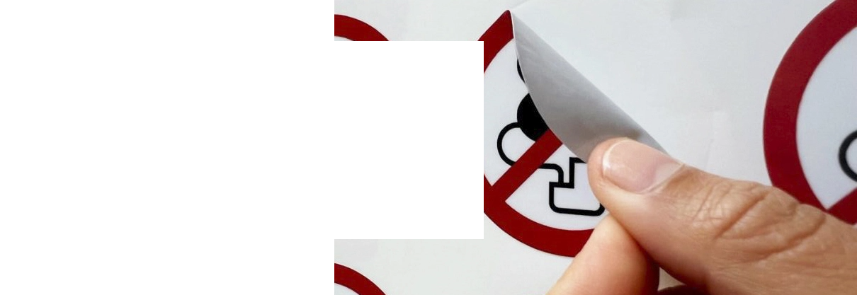 Photo avec un gros plan d’une main en train de coller une étiquette adhésive ronde de signalisation de la catégorie interdiction sur une surface blanche, plane et verticale 